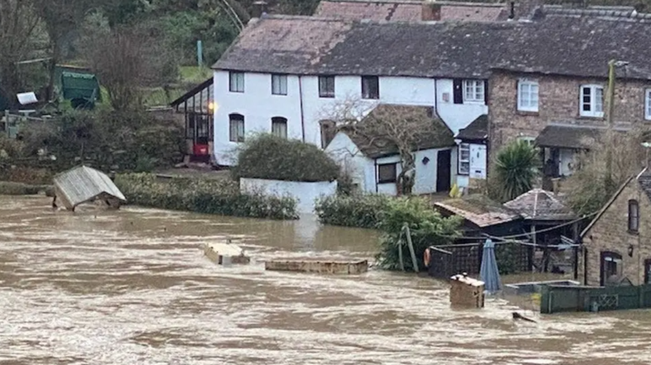 A row of white and brown terraced houses with brown flood water in some of the gardens and furniture floating downstream