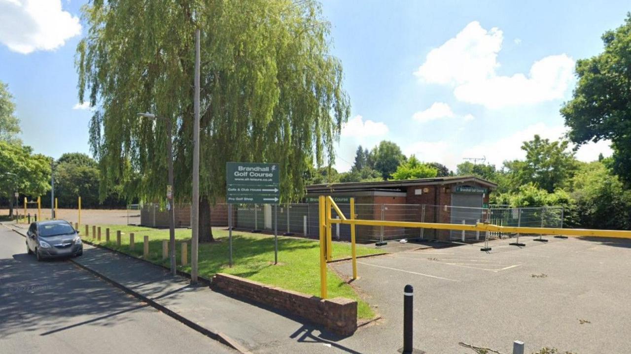 A building set back from the road, with grass and a Brandhall Golf Course sign between the building and the street. The building has been fenced off.