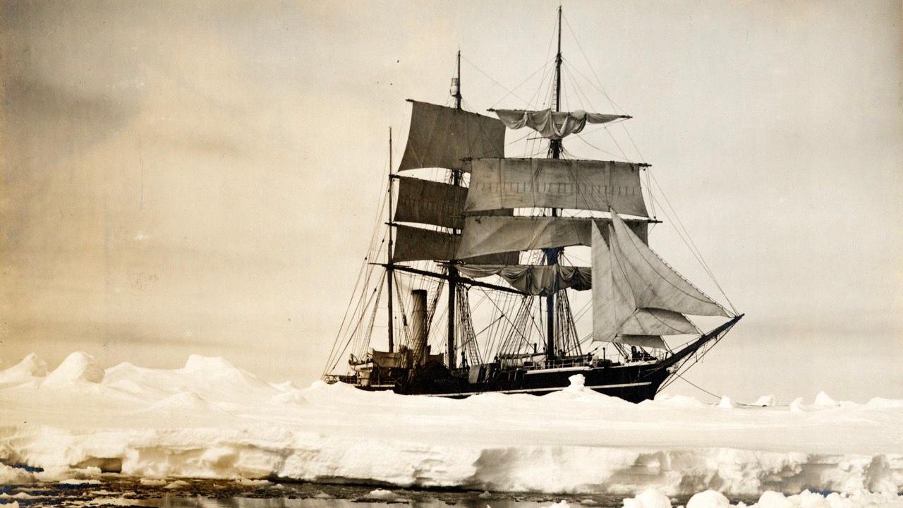 Black and white image of the Terra Nova ship sailing in Antarctica. The ship is wooden, it's sails are open. It is moving across ice-covered ocean.