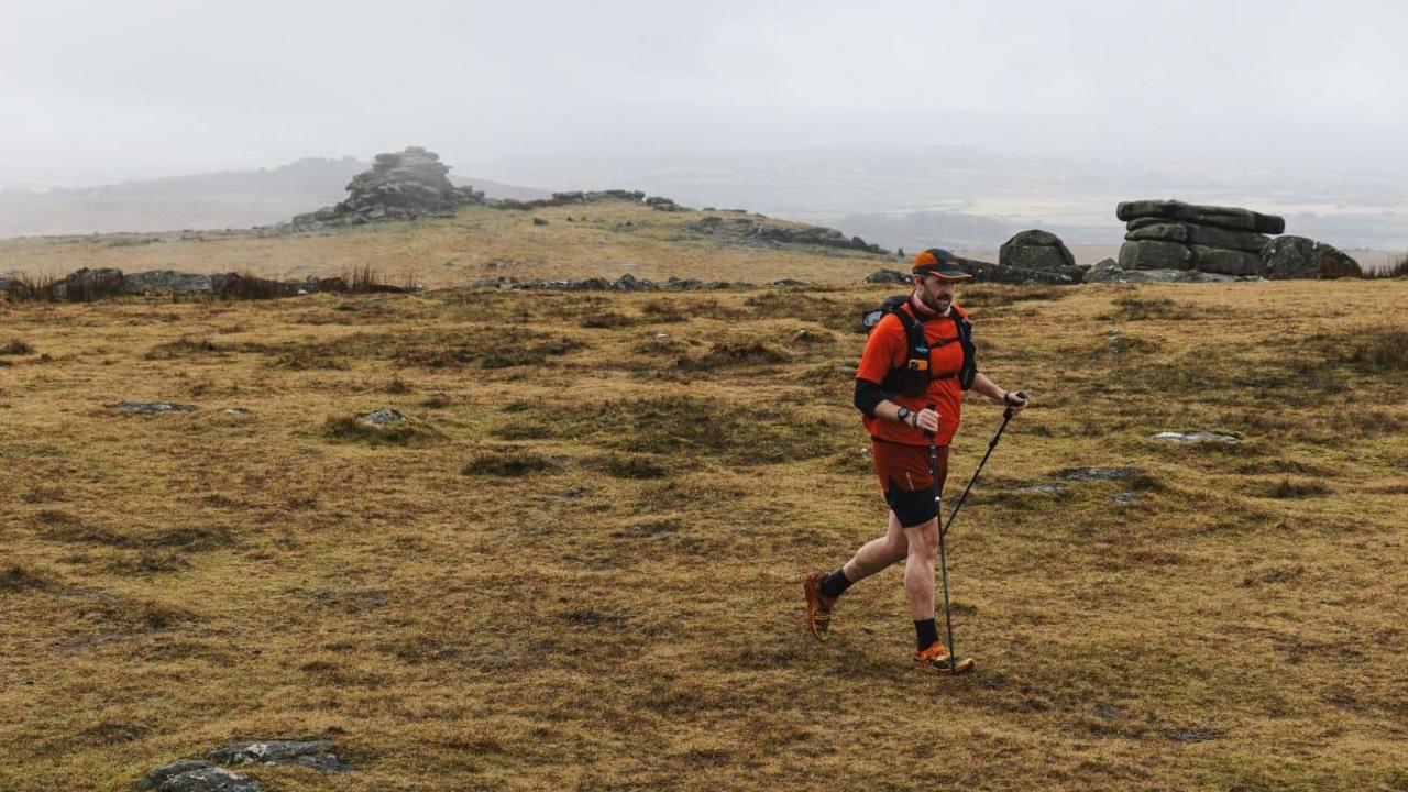 George Bell-Starr walking across a bare rocky heathland with the aid of Nordic poles. He is wearing an orange t-shirt and shorts, hiking boots, a hydration vest and black baseball cap.