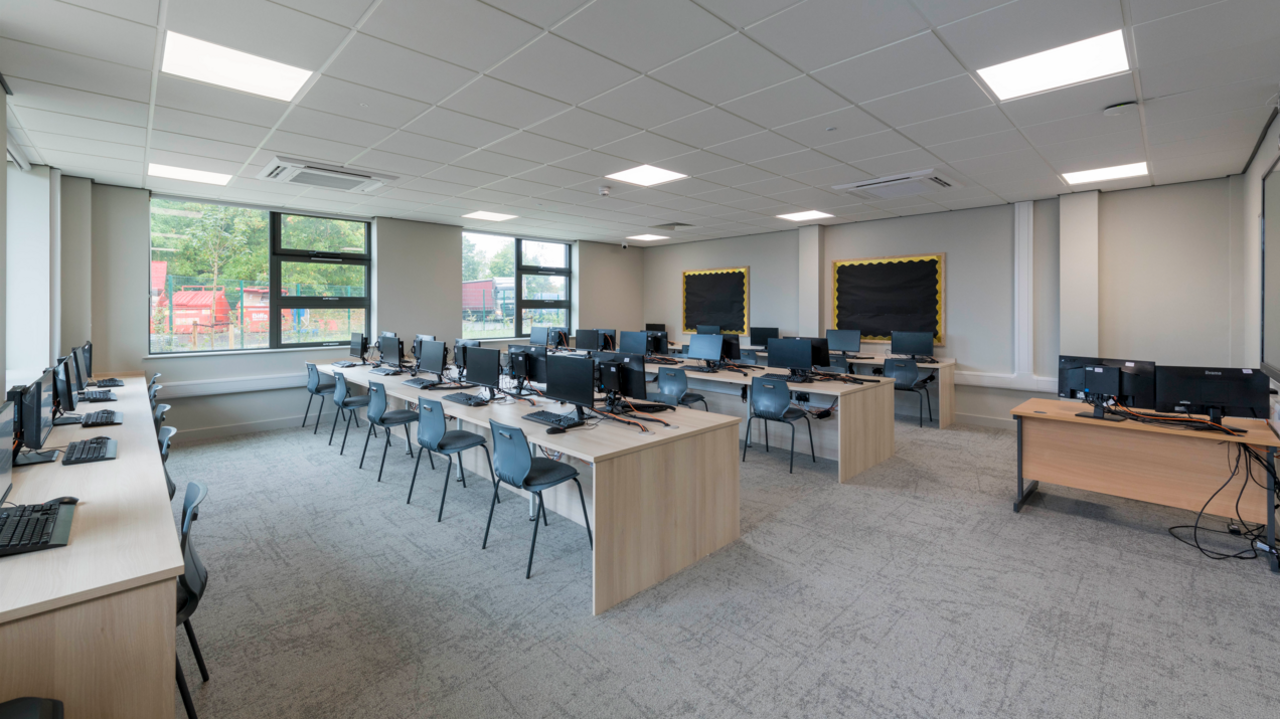 One of the newly developed IT classrooms. There are four rows of wooden desks with black computers and keyboards on top. The carpet in the room is a light grey and the wall and ceiling are white. 