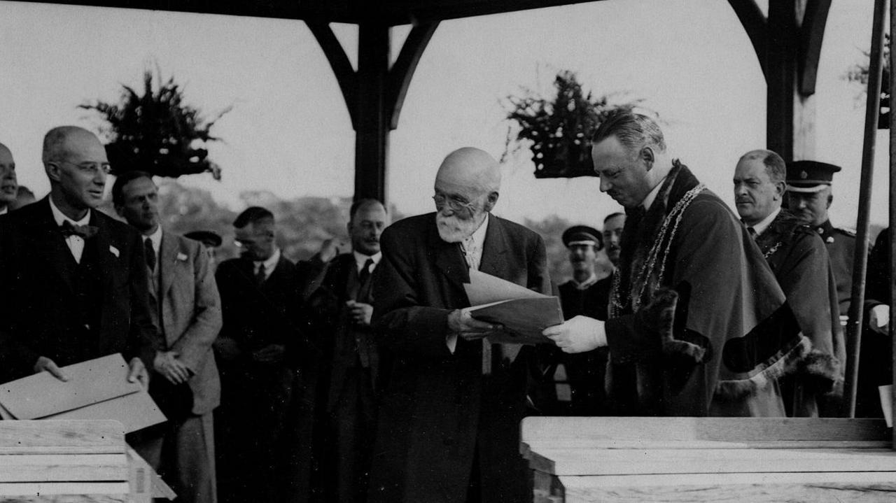 A group of men in suits stand in a bandstand. On the right a man in Lord Mayor's robes hands papers to Joseph Rowntree, a man with small round glasses and a white beard.