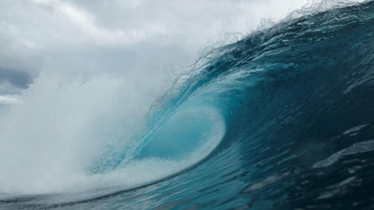 The crescent of a wave is seen crashing over. The wave is dark blue and lighter towards the tip of the wave and there is white spray coming away from the wave. The sky is cloudy.