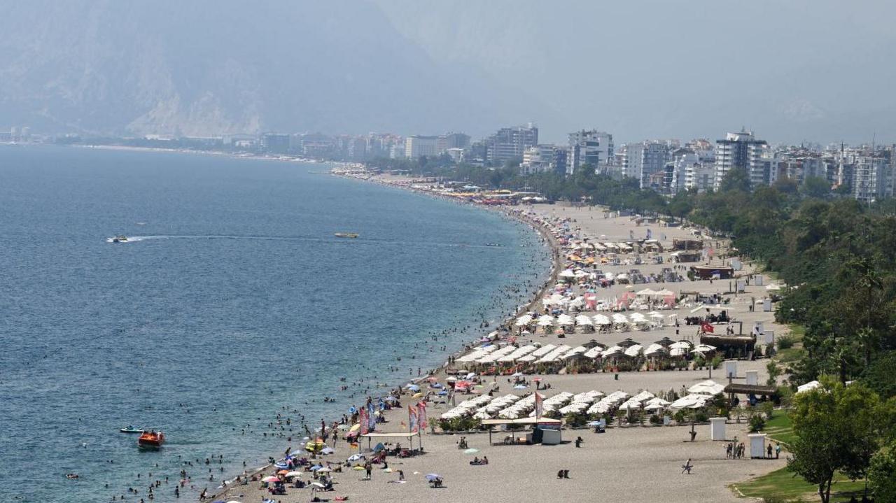 A picture taken from high up of a beach in Antalya, Turkey. To the left is the blue water, in the middle is the sand covered with people and umbrellas and to the right are high-rise buildings and trees.