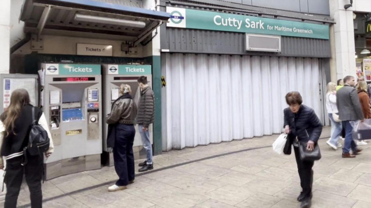 Exterior of Cutty Sark station with shutters pulled across and members of the public walking past