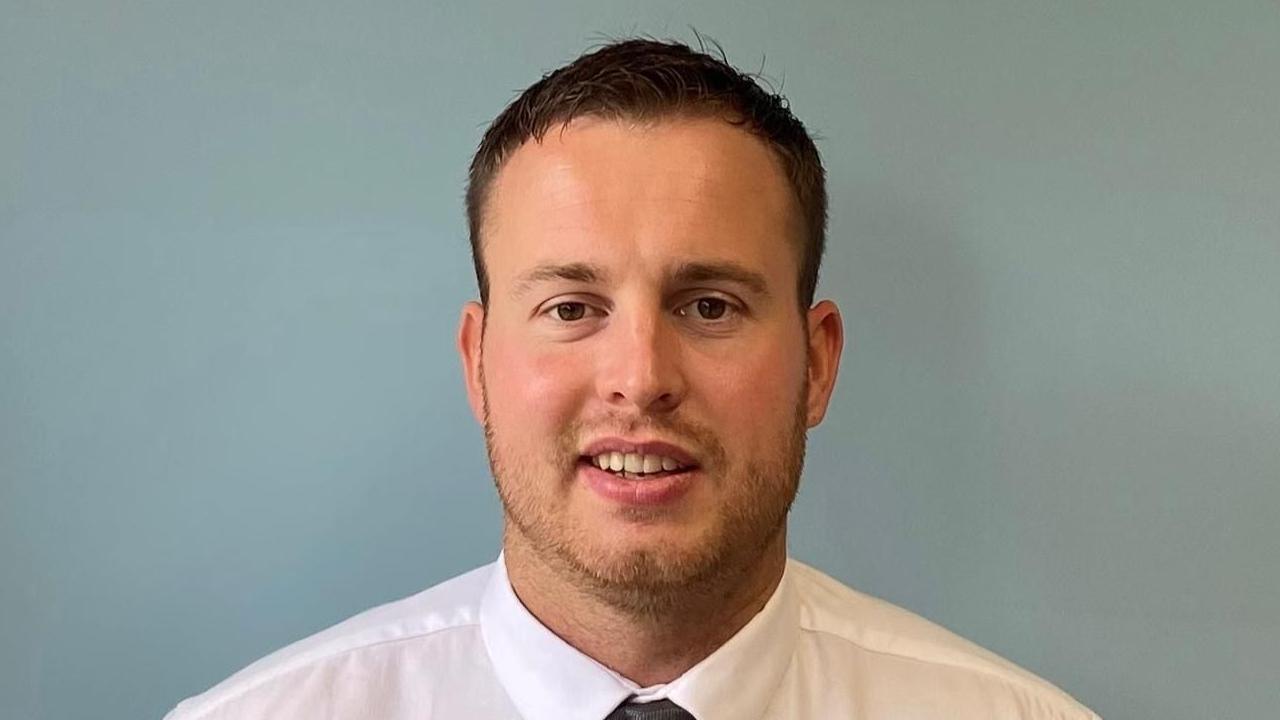 Ben Jackson has brown eyes, short, dark hair and a neatly trimmed short brown beard. He is wearing a white shirt and a black tie
