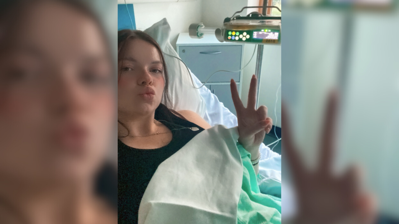 Verity Pitts lies in a hospital bed connected to a bedside monitor by a drip. She partly lies beneath a blanket while taking a selfie, raising the index and middle fingers of her left hand to form a 'peace' sign.
