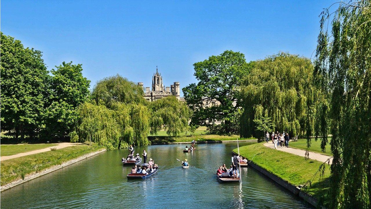 Boats on a canal with green paths on each side, a building rising above trees in the background and blue sky above 