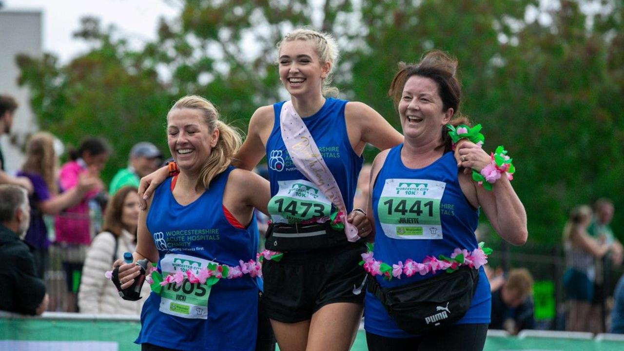 Three women who took part in 2022 and are wearing blue are arm in arm and smiling at the camera. Other people and trees in the background are blurred.