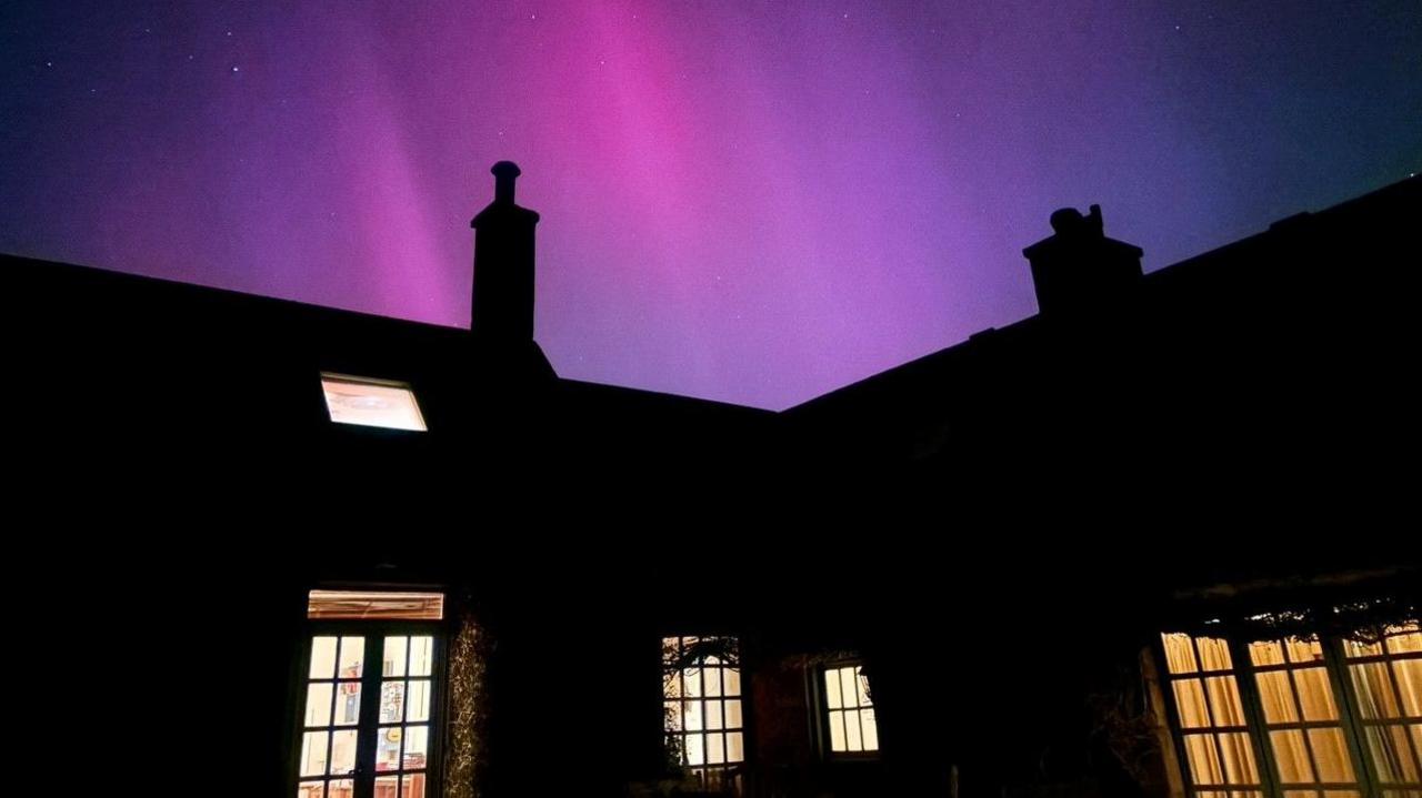 The blue and pink aurora appear to touch the roof of this house in Stenton, East Lothian. The house is one and a halft storeys tall and is in silhouette though its windows are lit brightly.