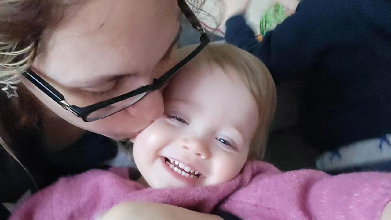 Emily and Isabelle Cooper. Emily is hugging her daughter and kissing her on her cheek as she smiles. Isabelle is wearing a pink jumper. Emily has black rimmed glasses.