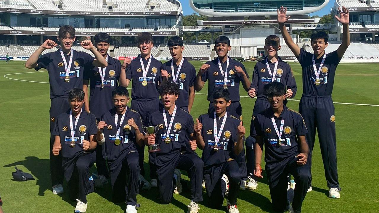 Twelve members of an U15 cricket team, dressed in navy, on the ground at Lord's. They are all wearing medals and have either thumbs up or fists raised. One member is carrying a cup.