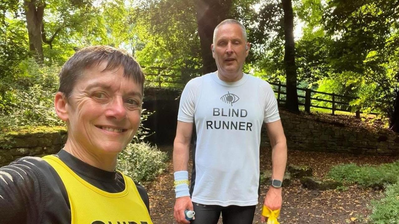 A selfie of Mr Goodall, who is wearing a white T-shirt which reads 'BLIND RUNNER' wearing a white wrist band on his right arm and an Apple watch on his left wrist. The image is being taken by Pascale Holden, a woman with short, dark-brown hair, wearing a black T-shirt and yellow vest. They are standing in a wooded area with a brick wall behind them. 