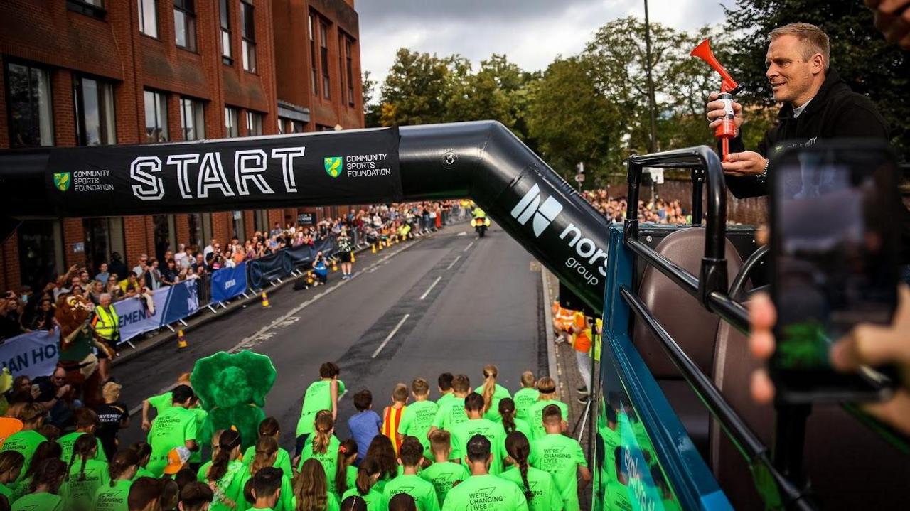 Norwich City FC Head Coach Johannes Hoff Thorup stood on a double decker bus with a horn in his hand while junior runners begin crossing the start line