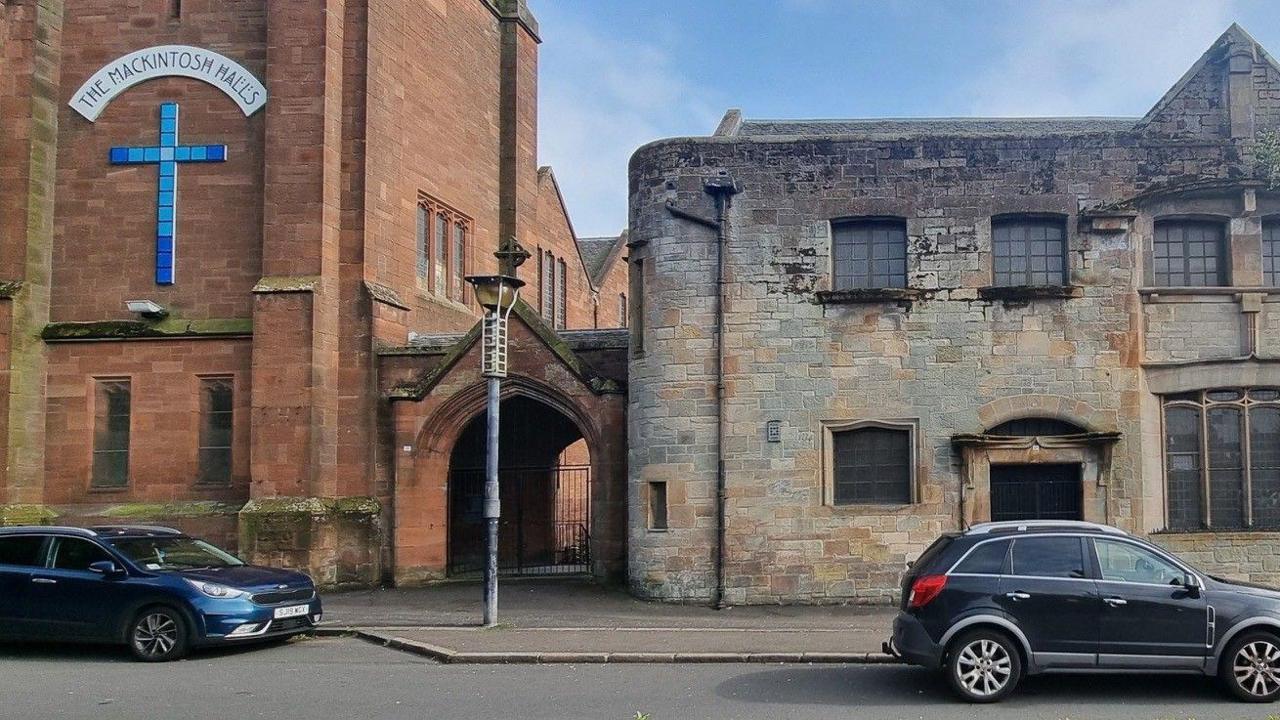 A church building with two cars parked in front of it. Part of the building is a red brick style, with a cross and The Mackintosh Halls on the wall.