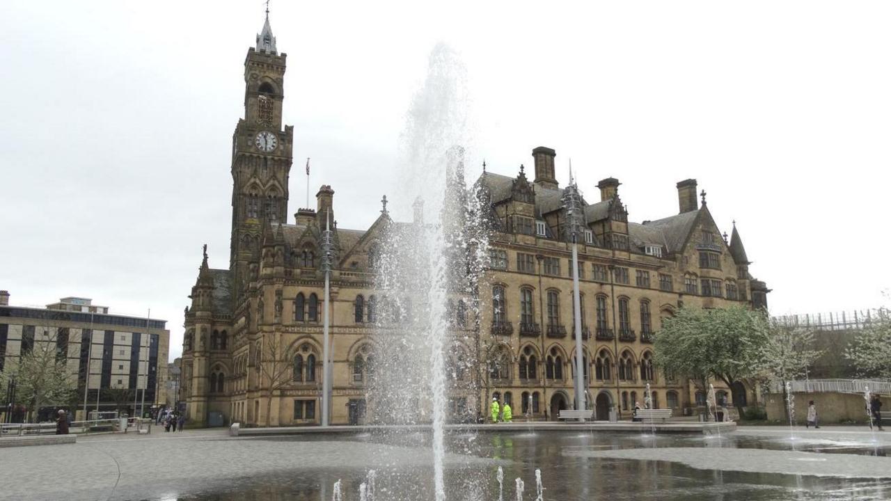 A large gothic town hall building with a tall clocktower. A fountain is in front of the building. A lower 1960s style building is to the left of the building.