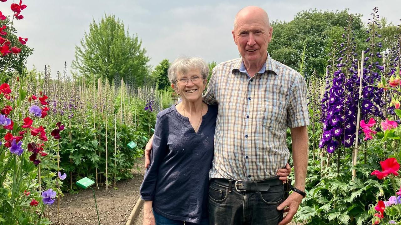 A couple stands in the middle of a path with their arms around each other. On either side, flowerbeds are filled with colourful plants.