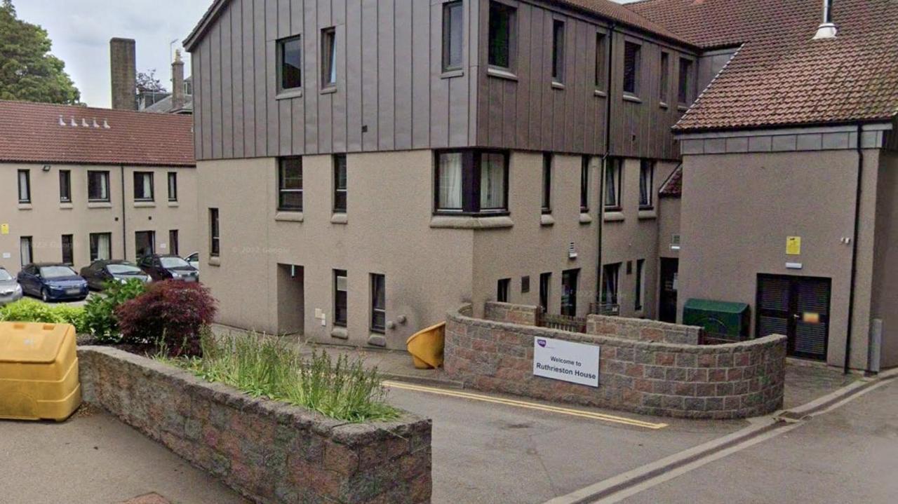 Ruthrieston House in Aberdeen behind a small brick wall with a white sign with black writing which says "welcome to Ruthrieston House", and included a purple VSA logo with white writing, The building has several windows, and a sloped roof. In the background of the picture is a secondary care home building.
In the foreground of the picture is another wall with some greenery inside it, next to a yellow salt bin.