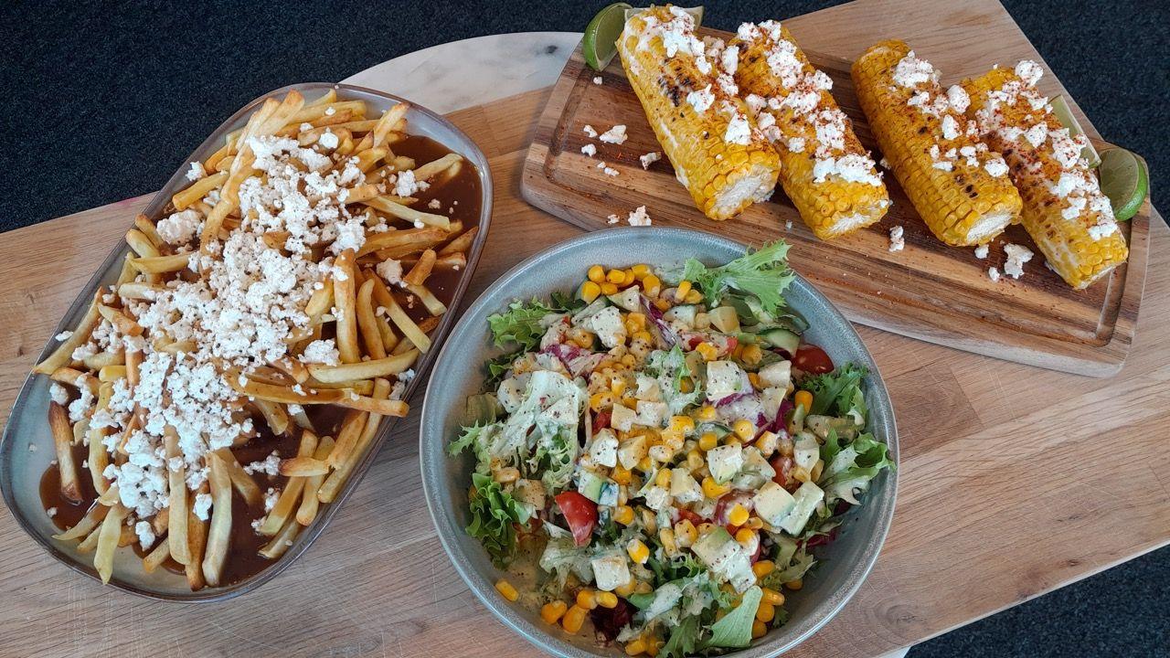 Ranch dressing in a bowl in the centre of the image, poutine on the left on a plate and corn on the cob on the right, all displayed on a wood slab. 