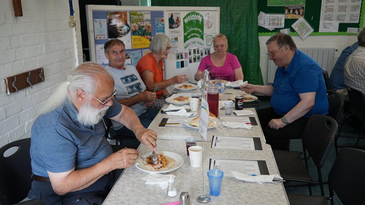 People sitting on plastic chairs around a white trestle table, eating food from white plates with knives and forks. 