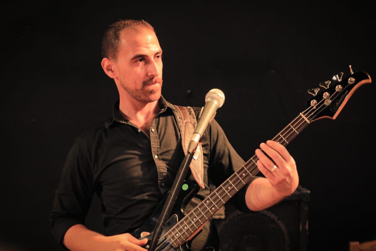 Guitar teacher Khamis Abu Shaban, in a black shirt, playing an electric guitar on stage - lit from the front with a dark background behind.