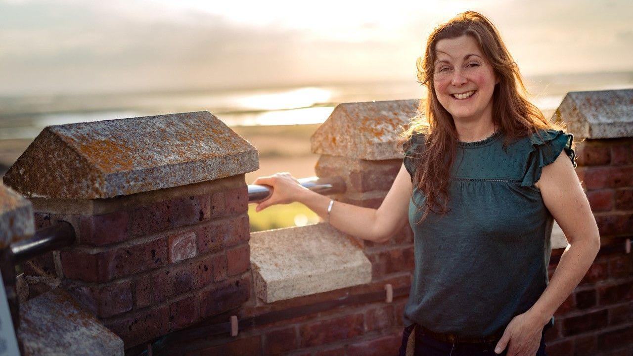 Michelle Nye-Browne standing on top of the Naze Tower. She has brown hair and is wearing a sleeveless top and holding on to a hand rail while smiling at the camera. In the background is a blurred view of marshes and water.