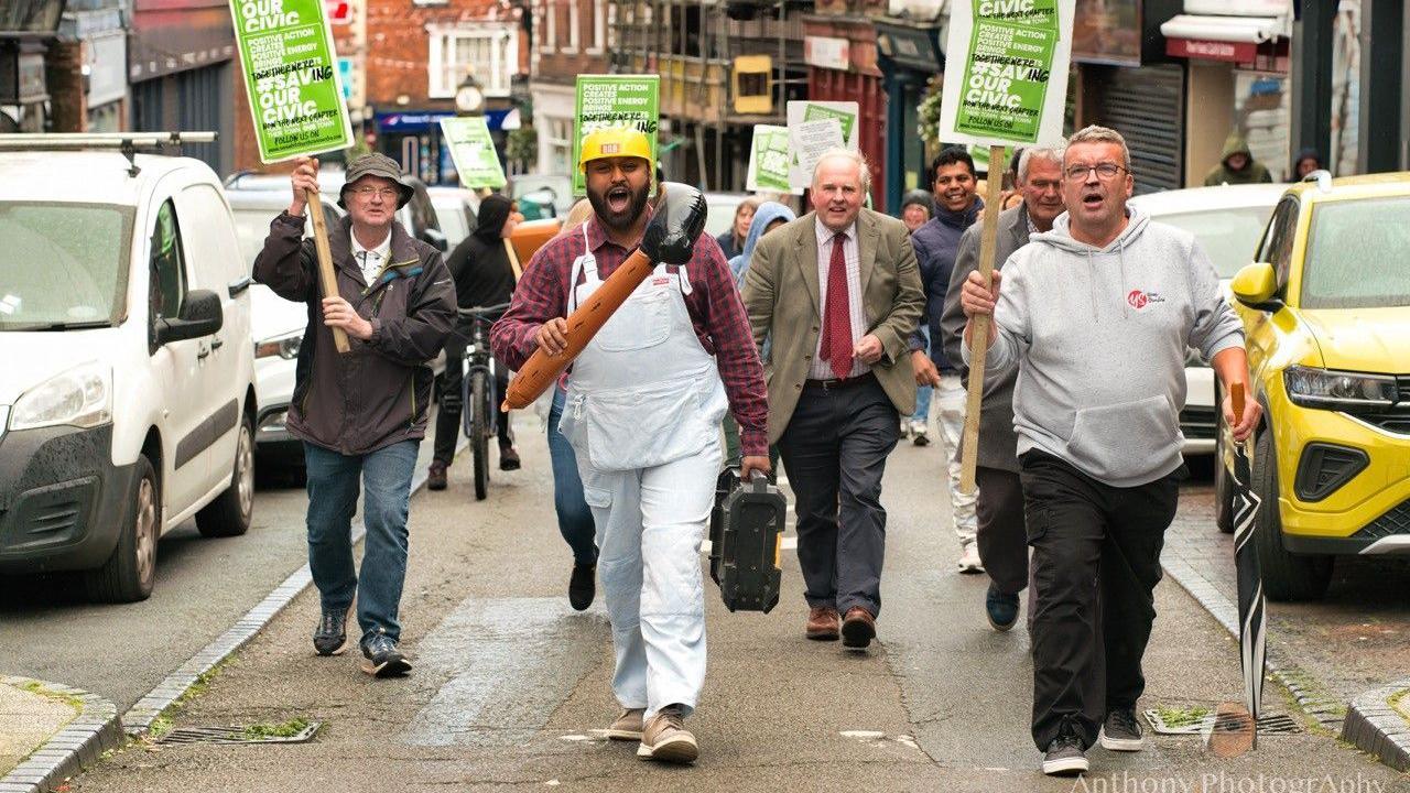 A group of people, one of them with an inflatable hammer, marching down a street. They are carrying green and white placards bearing the slogan "Save our Civic".