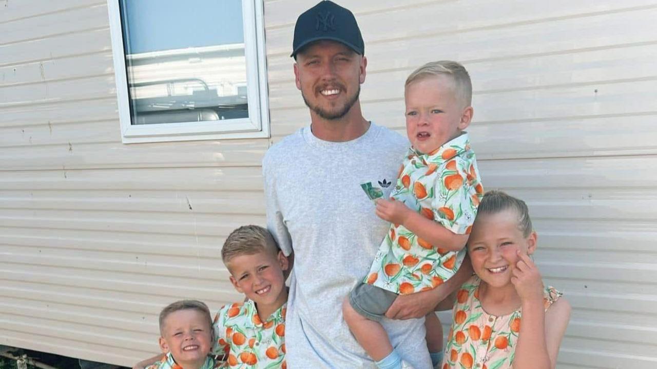 Nathan Osman standing in front of what appears to be a static caravan. He has a pale grey Adidas T-shirt on with a black New York Yankees logo. He is holding a young blonde-haired boy in his left arm and two his right there are two other young boys, both blonde. To his left is a young girl who is also blonde. All the children are wearing matching pale green shirts covered in oranges. The girl has the same pattern on her dress, which is pale pink