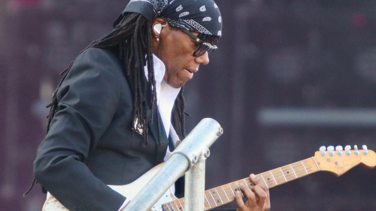 Nile Rodgers plays his guitar with ear phones in wearing suit and bandana