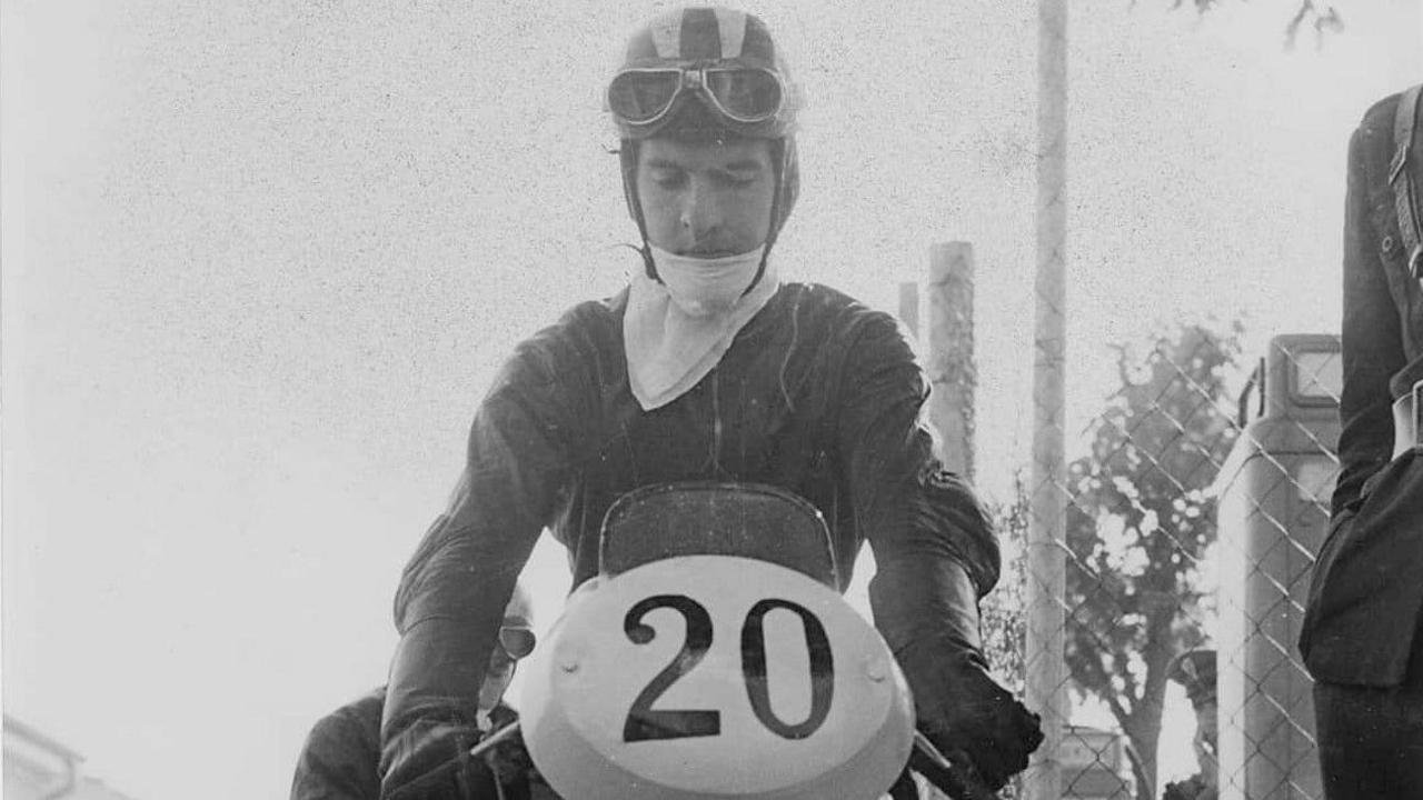 A black and white image of a man sitting on a motorbike with the number 20 on the front, he is wearing a white bandana around his chin and neck, a pair of goggles are sitting atop his head.