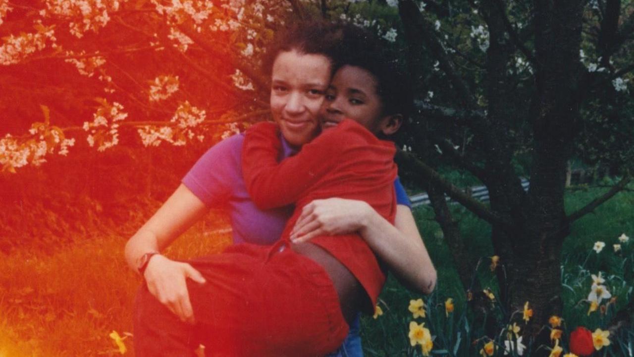 An older child carries a younger child in a flowering garden, surrounded by blossom-covered trees and spring flowers.  Warm red and orange light leaks across the image, giving the photograph a nostalgic, film-like atmosphere.
