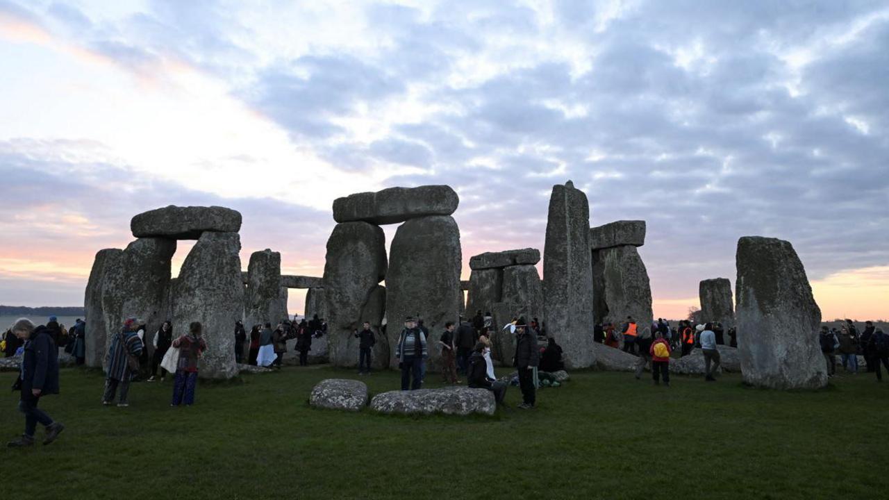 People gathered around the Stonehenge as the sky turns a pinky orange. There is a light layer of cloud.
