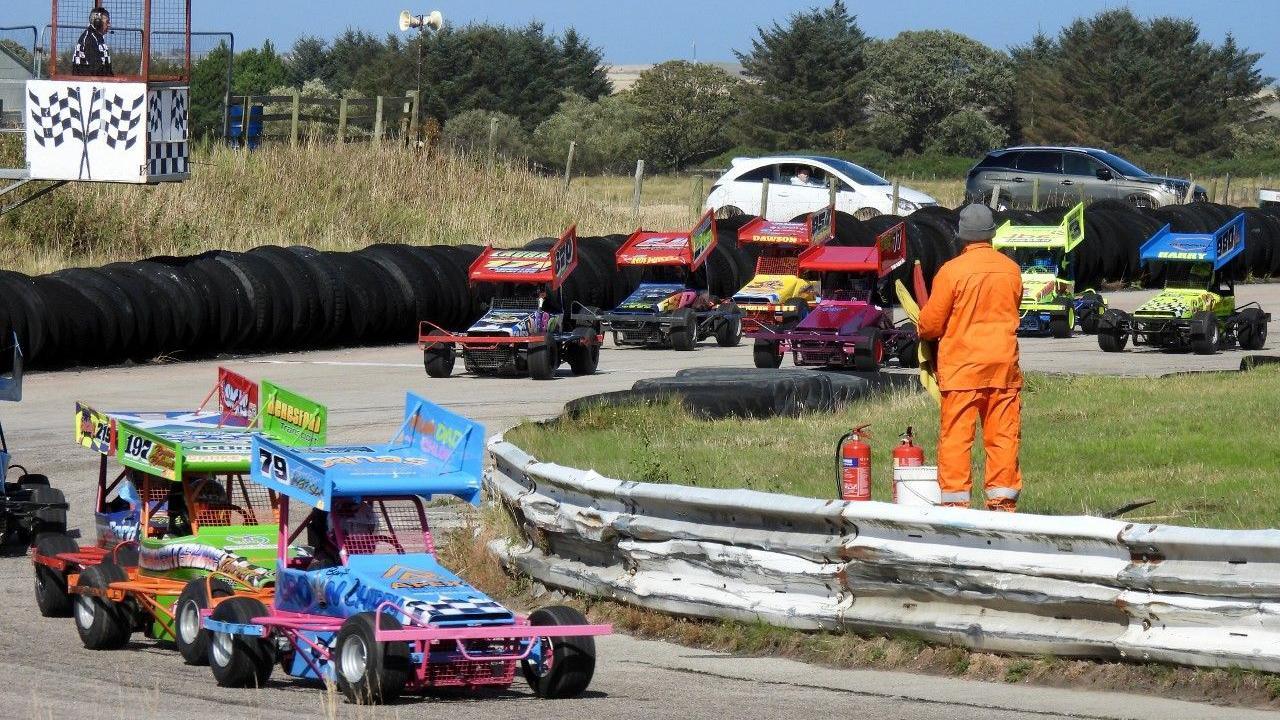 Brightly coloured small stock cars race each other on a track. There is a man in orange overalls in the centre with more small cars in the background.