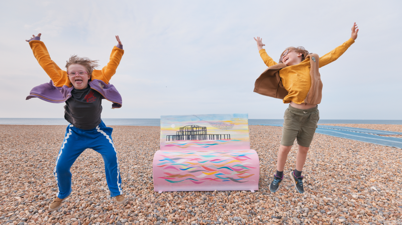 Two children jumping in the air on Brighton's shingle beach in front of one of the book bench sculptures which depicts the city's burnt out west pier. The bench is pink with multi coloured waves