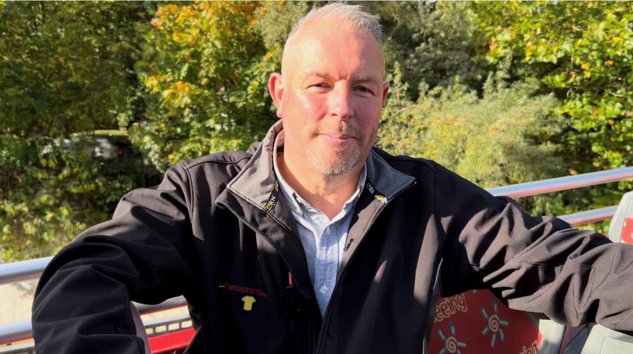 Chris Long-Pool sits on the top deck of an open-top bus in the sunshine. He has short grey hair and is wearing a dark coloured coat