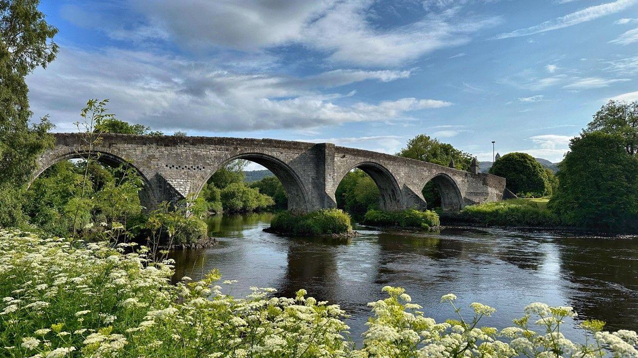 Viaduct with river flowing underneath, green trees and plants surrounding. Blue sky with clouds
