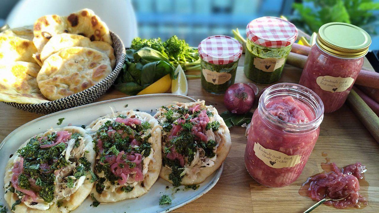 A plate of three flatbreads topped with pickled rhubarb and chicken, on a wooden counter beside a basket of flatbreads, herbs and lemons, and jars of homemade herb dressing and pickled rhubarb. One of the jars of pickled rhubarb is open and a spoon lies beside it with rhubarb pickle overflowing.