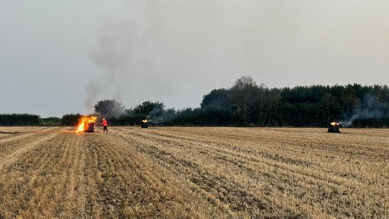 Bales can be seen on fire in a field. A firefighter is in the distance working to put out one of three fires in this photo.