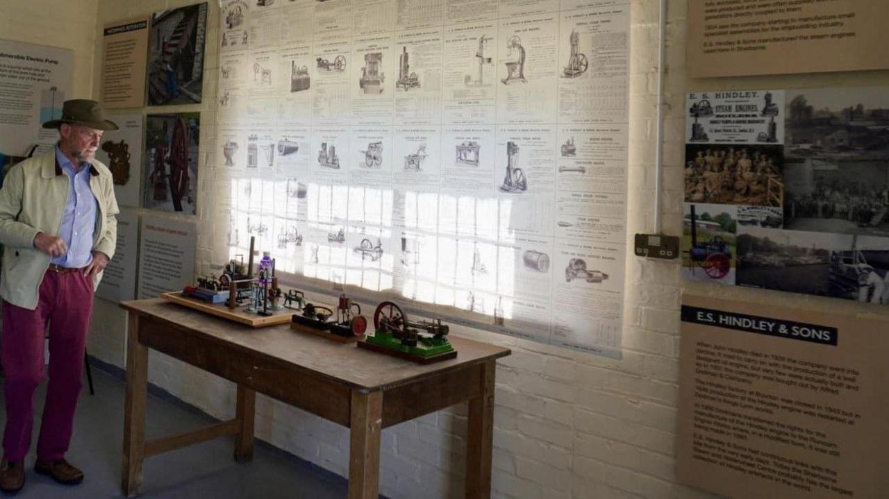 A man wearing a brimmed hat and red trousers looking at three models of pumping stations lined up on a display table. The walls of the room are covered with information boards.