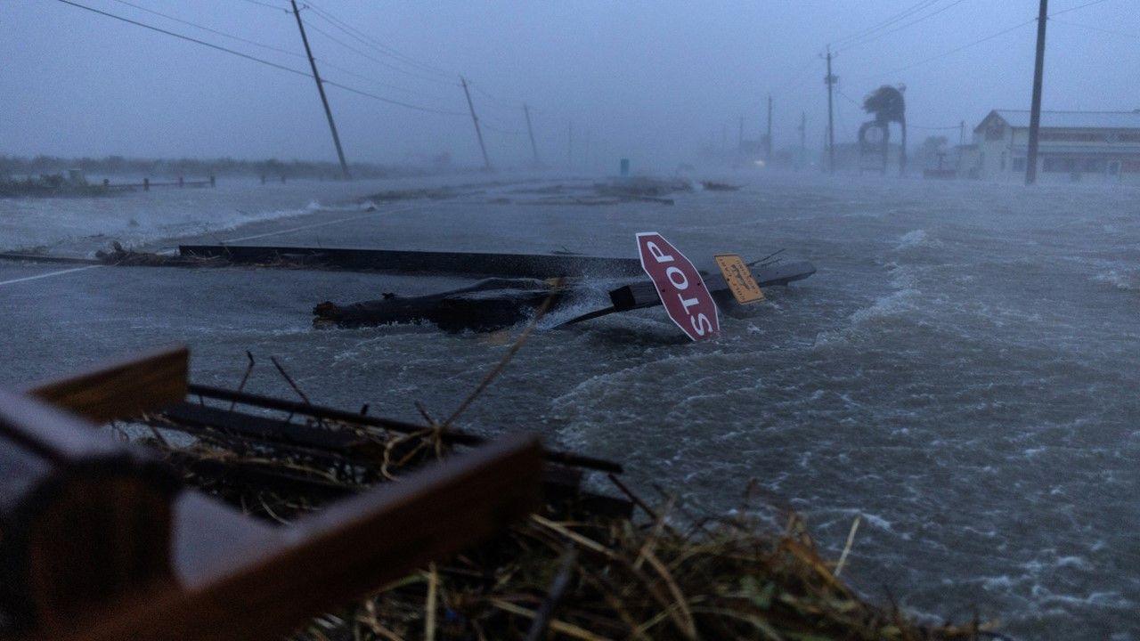 A road way is covered in water, fallen road signs and debris as trees are buffeted in the grey, rainy background