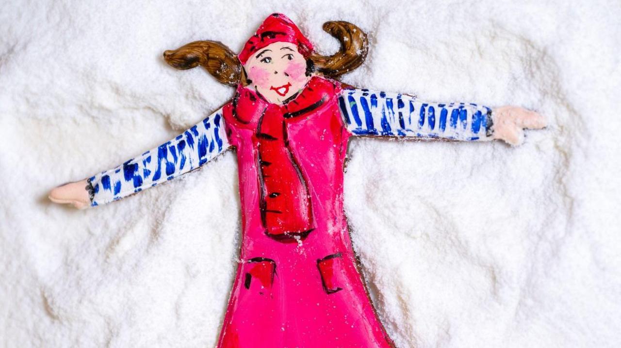 An iced biscuit model of a girl wearing a red hat with pigtails and a red scarf, coat and blue sleeves, laying on a white sugary surface.