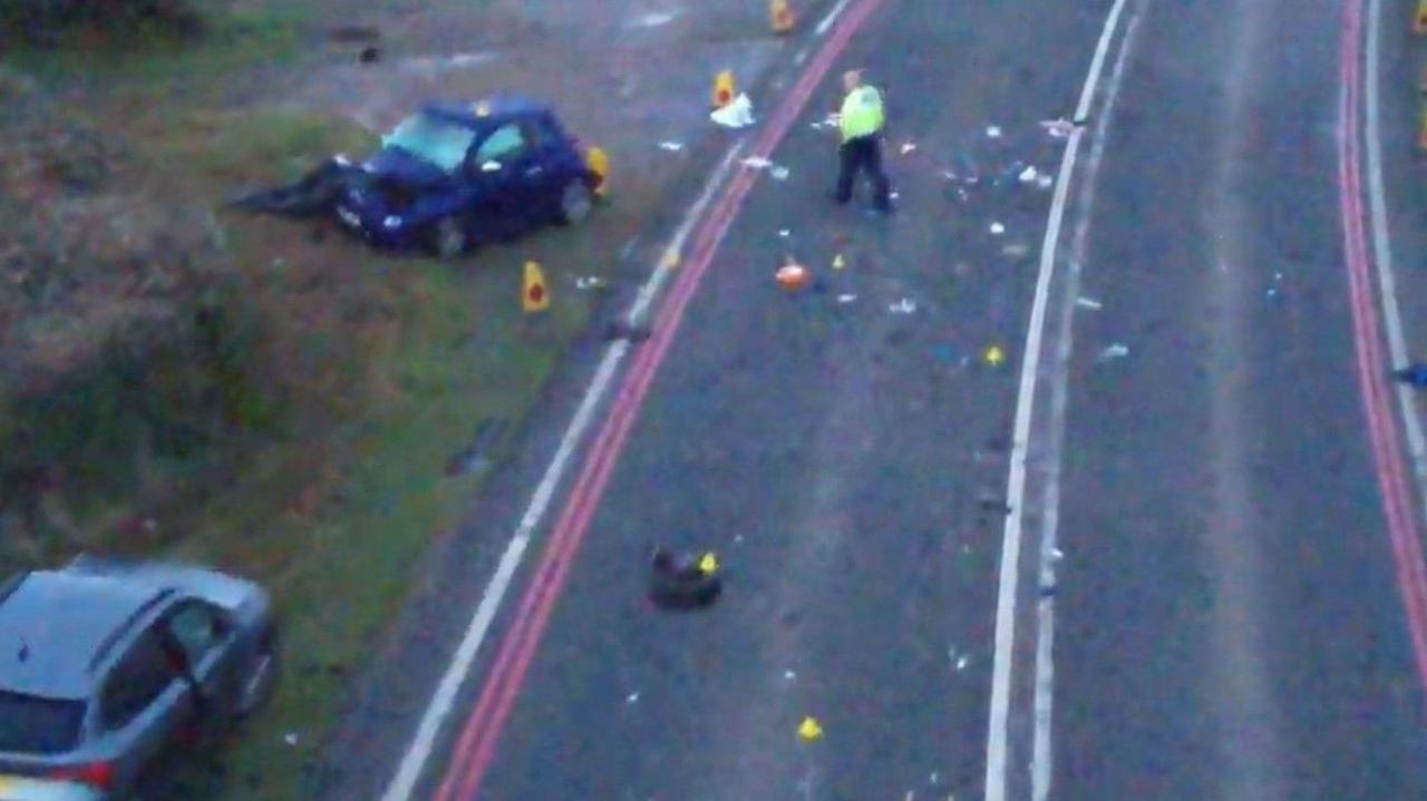 A still from drone footage of the collision scene on the A38 near Bristol. It shows debris on the road and a police officer walking towards a dark car which appears to be badly damaged