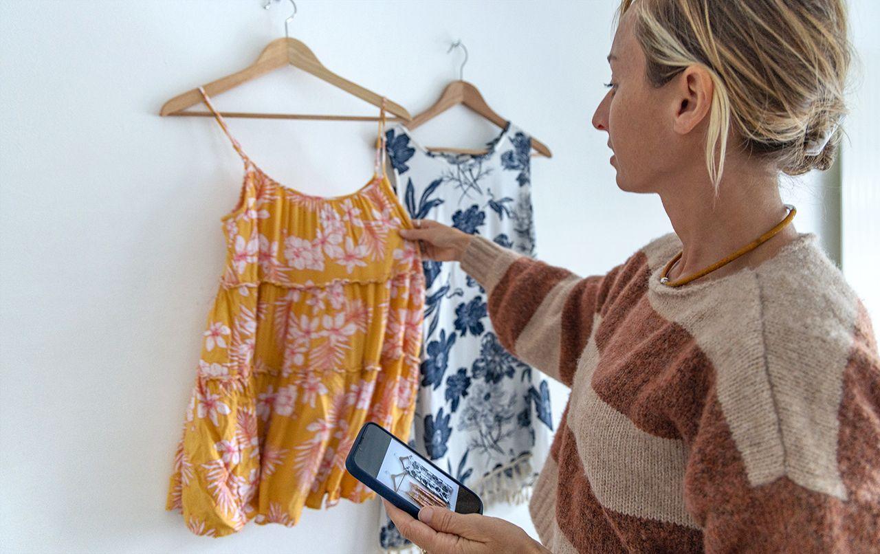 A woman adjusts a dress hanging on a wall in her home, while she holds her phone with the camera app open