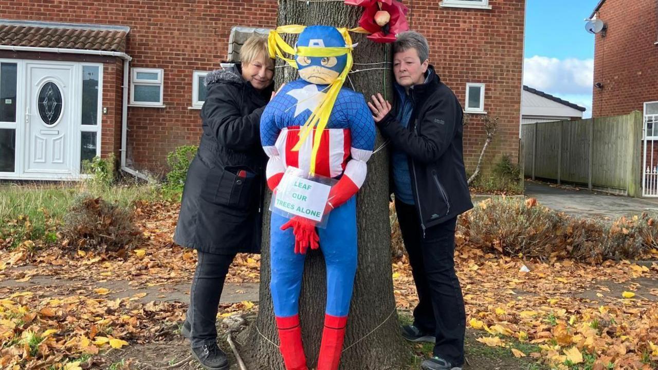 Two women stand on each side of a tree which has a large Captain America figure tied around the middle. The sign reads 'leaf our trees alone'.