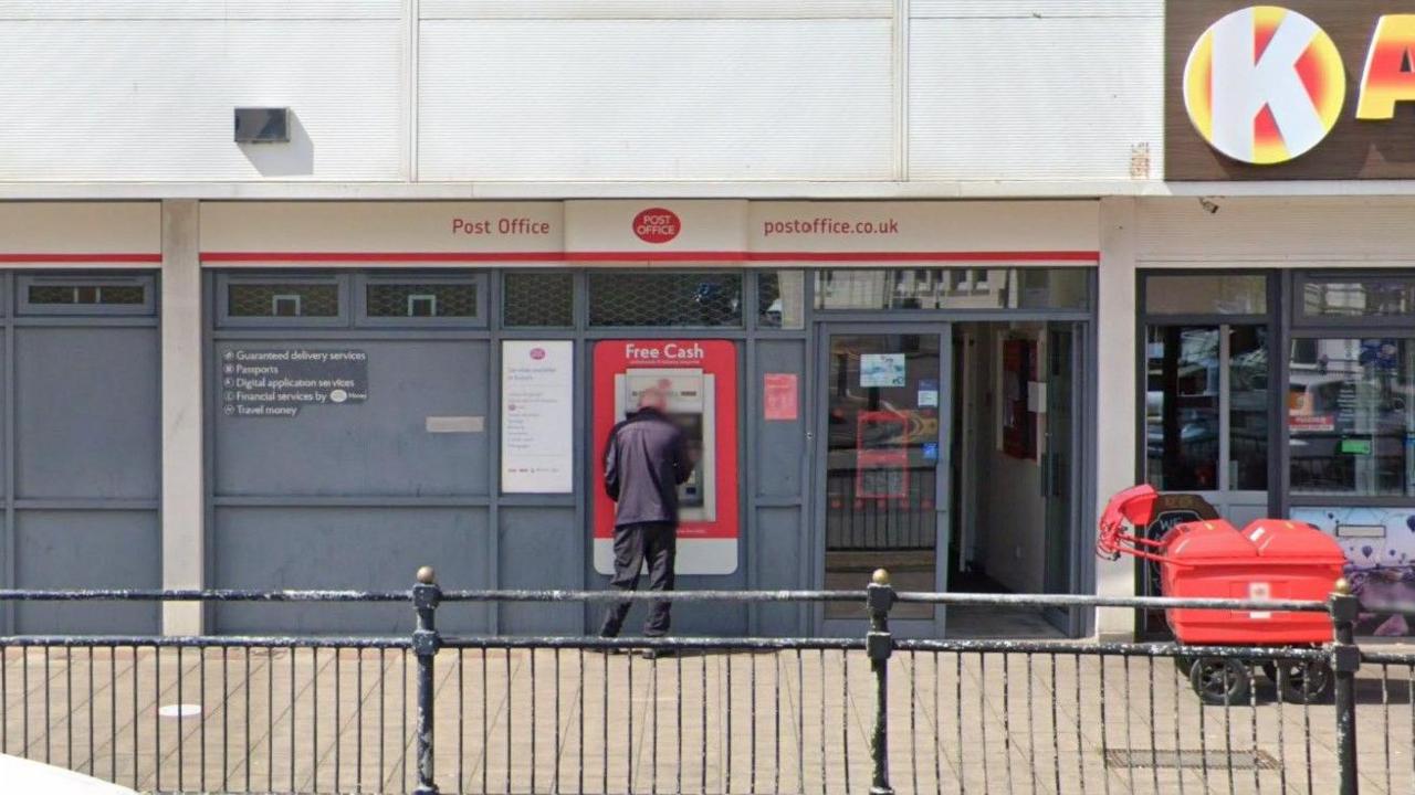 Man standing at cash point in front of the Post Office branch on a sunny day.