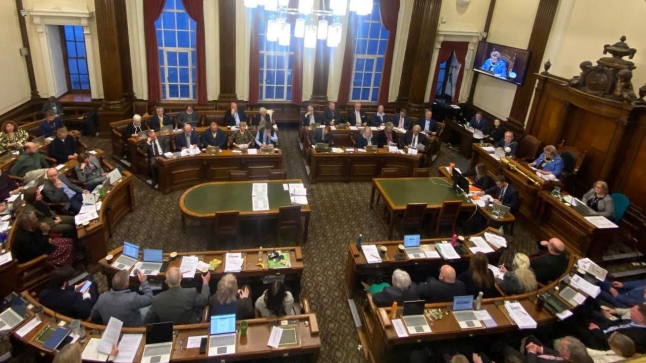 An internal view of Wirral Council meeting taken from above with rows of councillors sat at desks with papers and laptops in front of them.