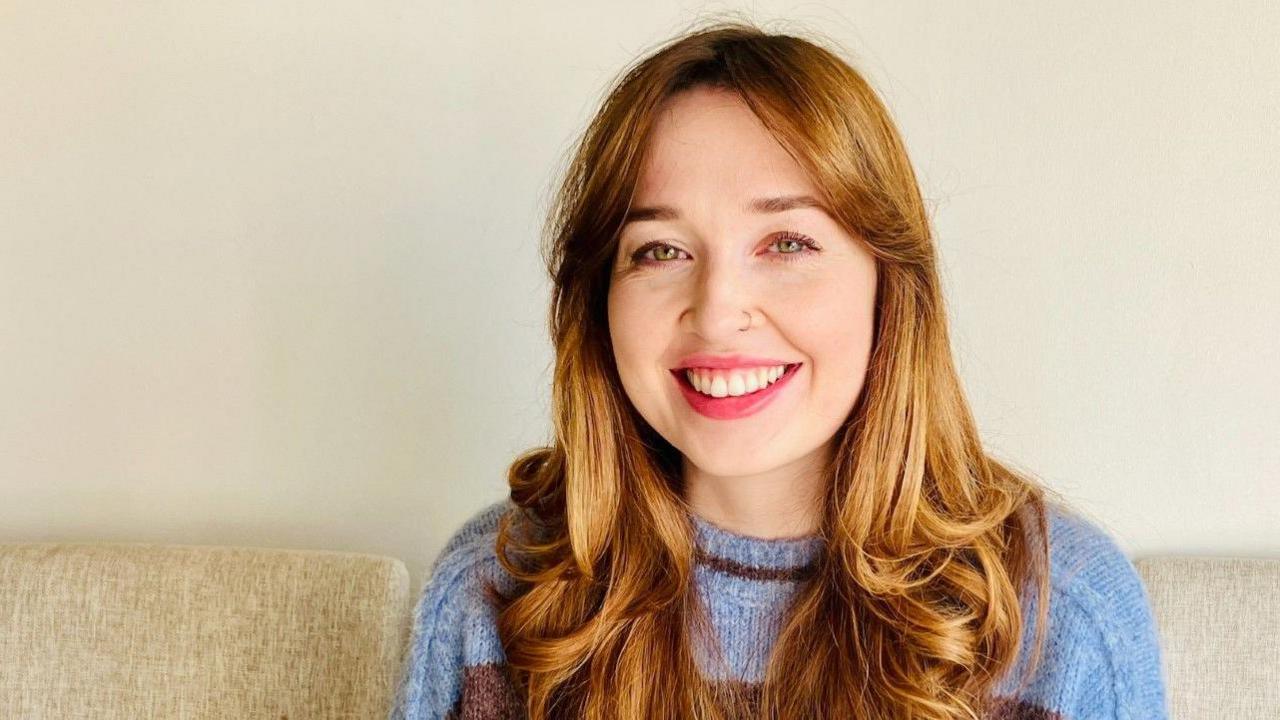 Kate Driver is smiling at the camera. She has slightly curled hair and is wearing a light blue and purple jumper.