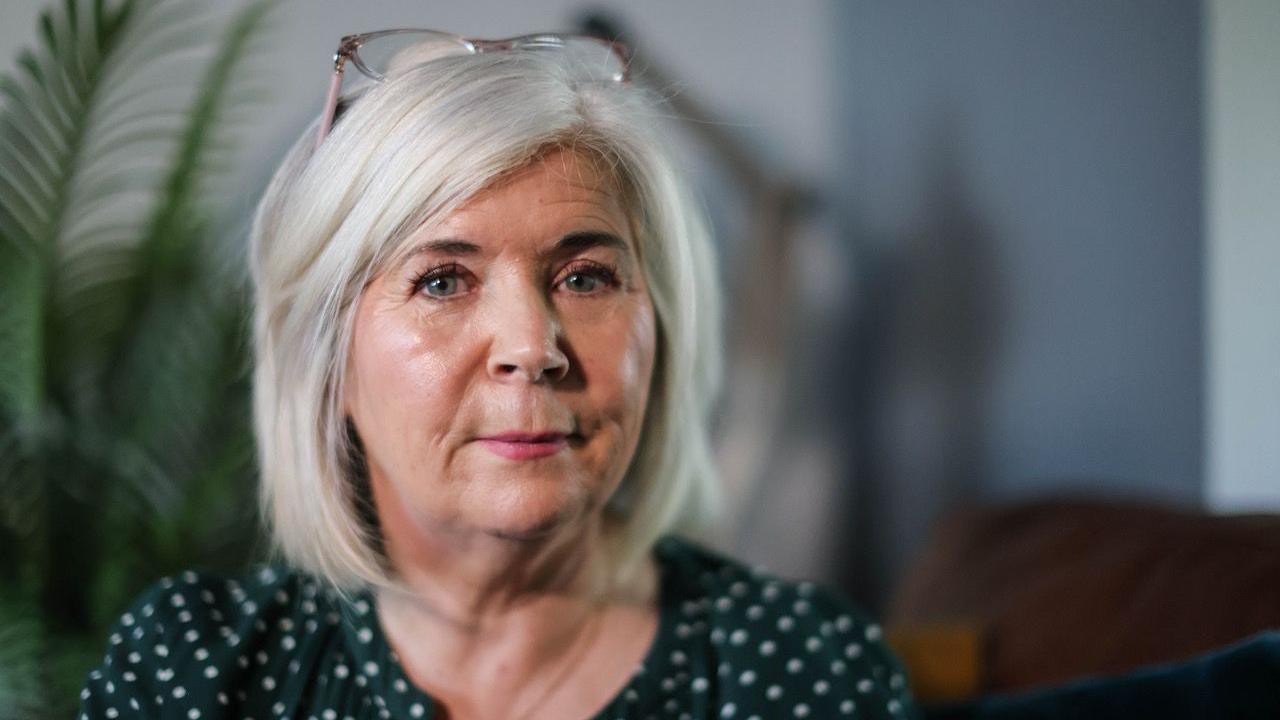 A 59-year-old woman wearing a black-and-white polka dot top looks at the camera