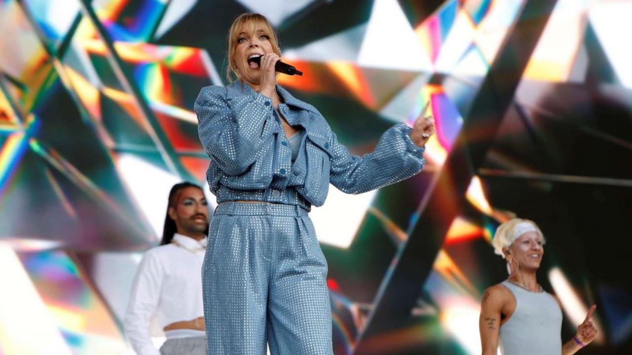 Becky Hill, wearing matching blue trousers and jacket. She is standing on a stage with two backing dancers in the background. Hill is holding a microphone and is holding her other arm out whilst singing.