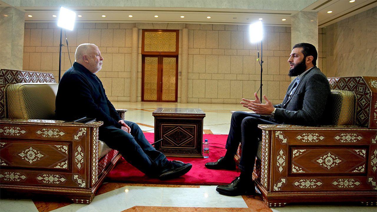 Jeremy Bowen and Ahmed al-Sharaa sit opposite each other during the interview, both sitting in armchairs in a large room with a red carpet on the floor. 